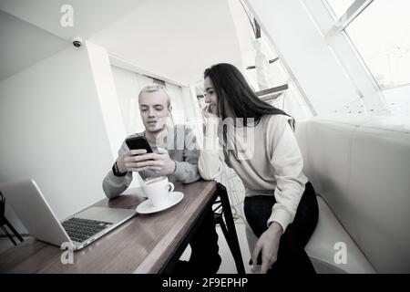 Young couple meets in coffeehouse. Blonde man showing something fun in his smartphone to brunette attractive woman. Modern laptop on table. Social Stock Photo