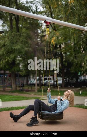 A Vertical shot of a blonde cheerful woman having fun on the swing in the park Stock Photo