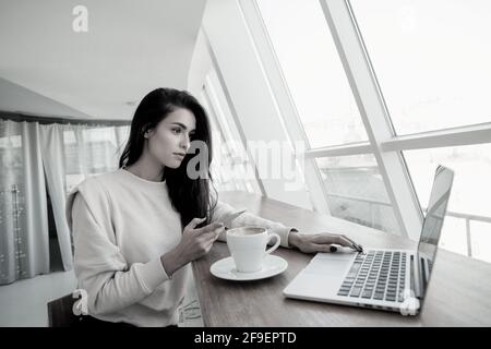Freelancer woman working on laptop and typing on smartphone to customer. White room on background. Woman programmer looking for a bug in code and Stock Photo