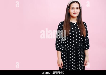 Portrairt of a modest-looking young woman in a polka dot dress over pink background. She's subtly smiling, with headband on her head and long straight Stock Photo