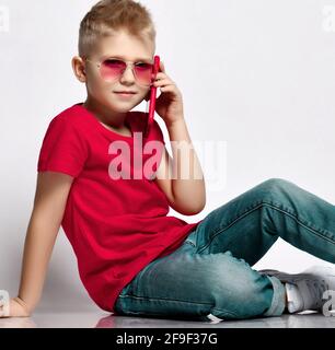 Stylish kid boy child in red t-shirt, jeans, sneakers and sunglasses sitting on floor talking on smartphone Stock Photo