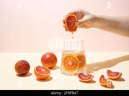 Female hand squeezing half of ripe juicy blood orange into a glass of functional water with citrus slices on a yellow-pink background in bright light. Stock Photo