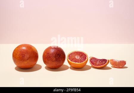 A row of whole and cut into halves and slices of blood oranges on a yellow-pink background in bright light, close up. Stock Photo
