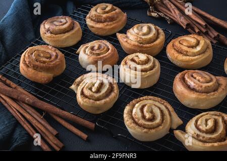 Homemade cinnamon rolls on a cooling rack with cinnamon sticks and dark blue cloth. Stock Photo