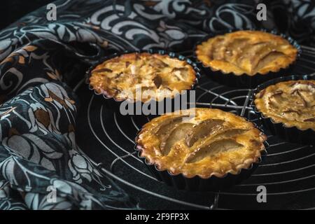 Baked apple tartlets in black tart dishes on a cooling rack with black and white patterned cloth as background. Stock Photo