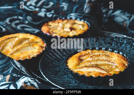 Three baked apple tartlets in black tart dishes on a black plate with black and white patterned cloth as background. Stock Photo