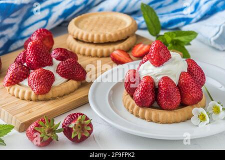 Strawberry tartlet on white plate, decorated with blossoms and mint leaves, in the background wooden board with tartlets and strawberries and blue and Stock Photo