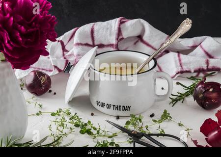 Potato soup in white bowl with handle and lid on white table with black background, decorated with red onions, vintage scissors, purple peopny blossom Stock Photo