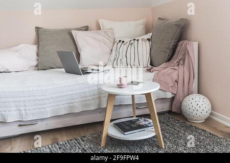 Cozy room with bed, grey and rose pillows, coffee table, notebook, mobile phone and coffee cup prepared for a break, interior in scandinavian style Stock Photo