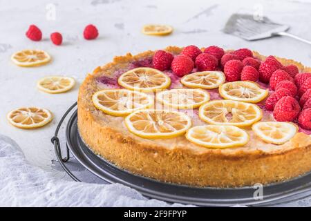 Cheese cake with raspberries and dried lemon slices on white background, some raspberries, lemon slices and cake server lying on the table. Closeup. Stock Photo