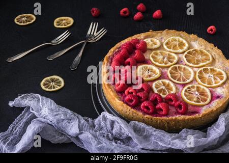 Cheese cake with raspberries, dried lemon slices, forks and lilac cloth on dark background Stock Photo