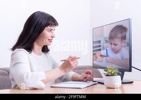 A female teacher sitting in front of a computer monitor holds a pen and teaches a small child to write. A small child is learning to write. Online lea Stock Photo