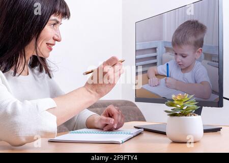 Attractive female teacher sitting in front of a computer monitor holds a pen and teaches a small child to write, close-up. A small child is learning t Stock Photo