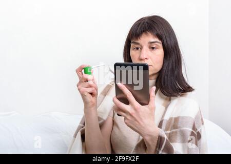 Portrait of a sick woman covered with a blanket holding a spray for treating a throat and communicating with a doctor on a tablet. Distance medicine c Stock Photo