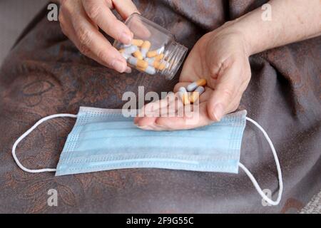 Elderly woman with pills in wrinkled hands on medical mask. Taking medication in capsules during coronavirus pandemic Stock Photo