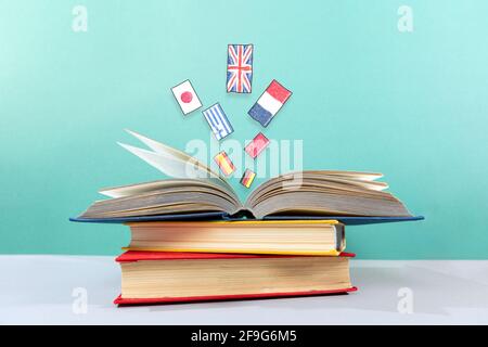 On the table is a stack of books, with an open book on top, from which flags fly out. Turquoise background. The concept of learning foreign languages. Stock Photo