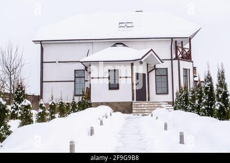 Residential two-storey country house. A house with roses in the shelter from the winter cold under the snow. The path to the house is cleared of snow. Stock Photo