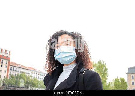 young black curly-haired woman smiling with mask on street with out-of-focus buildings in background Stock Photo