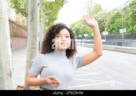 Woman with curly hair hailing a cab or uber with raised hand while holding a cell phone in the city. Horizontal photograph. Stock Photo