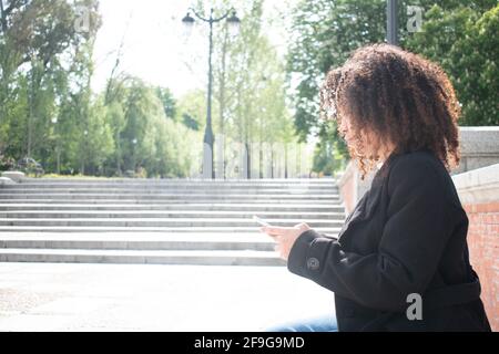 young woman with curls sitting on a bench using mobile phone outside with a background of stairs and a park. Stock Photo