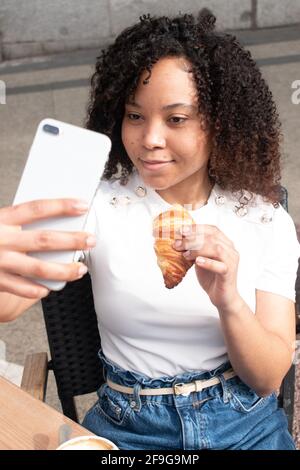 Young black woman with curls eating a croissant while taking a selfie outside a cafe. Stock Photo