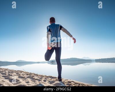 Athlete stretches his thigh muscles on the shore of the beach before running. Sunny morning training Stock Photo