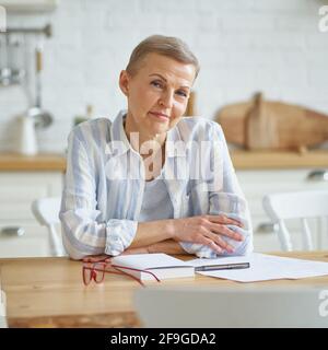 Home office. Portrait of beautiful middle aged woman smiling at camera while sitting at wooden table in kitchen and working with documents. Senior people and distance work concept Stock Photo