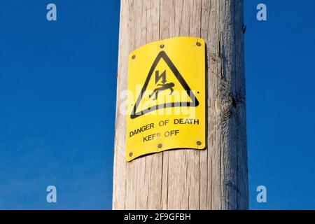 Close up of a yellow UK warning sign attached to a wooden electricity pylon warning of a danger of death from electrocution and to keep off. Stock Photo