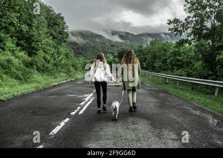 Sisters walking in the middle of an abandoned road in an apocalyptic landscape with a dog while holding hands Stock Photo