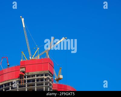 Tower cranes atop an unknown unfinished skyscraper against a clear blue sky during the day. The concept of the construction industry in the metropolis Stock Photo