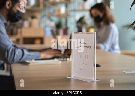Closeup of social distance sign standing on table with people working on pc. Stock Photo