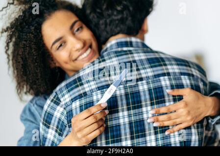 Expecting for a baby. Positive pregnancy test concept. Joyful african american young woman hugs her beloved husband, holding a positive pregnancy test, happy about good news, happy young family Stock Photo