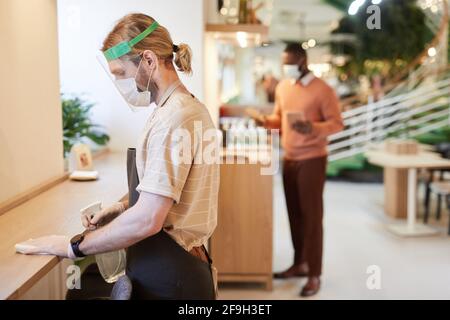 Side view portrait of male cafe worker wearing mask and face shield while sanitizing tables and cleaning furniture, copy space Stock Photo