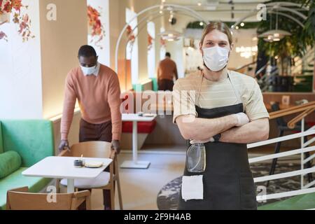 Portrait of young man wearing mask and looking at camera while posing with arms crossed in cafe interior and holding sanitizing supplies, copy space Stock Photo