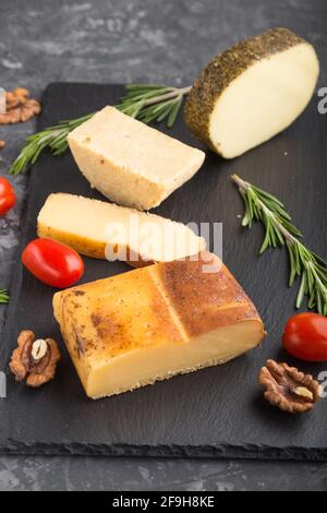 Smoked cheese and various types of cheese with rosemary and tomatoes on black slate board on a black concrete background. Side view, close up, selecti Stock Photo