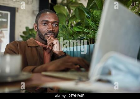 Portrait of modern African American man working with laptop while sitting at table in eco friendly green cafe interior, copy space Stock Photo