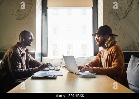 Side view portrait of two smiling business men using laptops while working at cafe table lit by sunlight in loft interior, copy space Stock Photo
