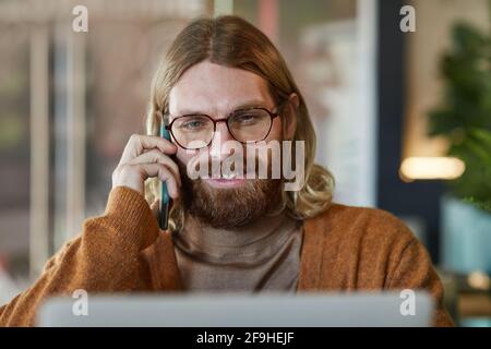 Front view portrait of contemporary bearded businessman speaking by smartphone and using laptop while working at cafe table Stock Photo