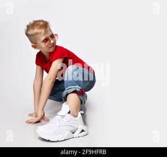 Active, frolic blonde kid boy in red t-shirt, blue jeans, white sneakers and sunglasses is in ready to run position Stock Photo