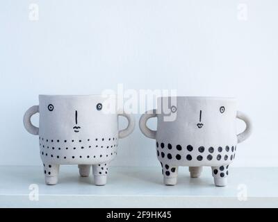 Two empty cute face ceramic plant pots on white wooden shelf isolated on white wall background with copy space. Small modern DIY cement planter trendy Stock Photo