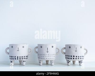 Three empty cute face ceramic plant pots on white wooden shelf isolated on white wall background with copy space. Small modern DIY cement planter tren Stock Photo