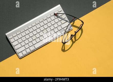 White keyboard and reading glasses on yellow and black background top. Flat lay. Stock Photo