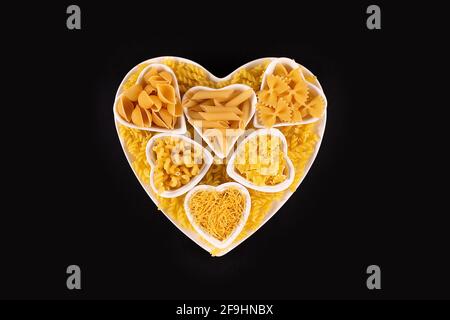 Pasta food selection in heart shaped porcelain dishes over black background. Uncooked, Dried pasta noodles. Love pasta concept Stock Photo