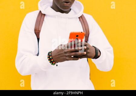 Young african black man against a yellow wall wearing a white sweatshirt and a backpack listening music on wireless headphones while looking at smart Stock Photo