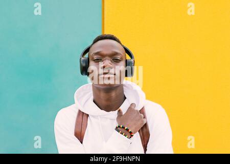 Young african black man against a yellow and green wall wearing a white sweatshirt and a backpack listening music on wireless headphones while looking Stock Photo