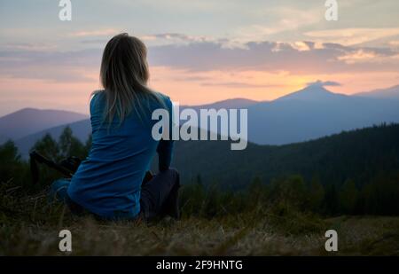 Back view of female hiker who resting after day of hike and watching at sunset above mountain beskids. Tourist enjoying landscape of mountain hills and evening sky when sun setting over horizon. Stock Photo