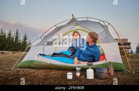 Young couple, waking up after night's sleep in tent outdoor in the mountains, happily smiling at each other, planning delicious breakfast. Pleasant morning of family pair in mountains campsite. Stock Photo