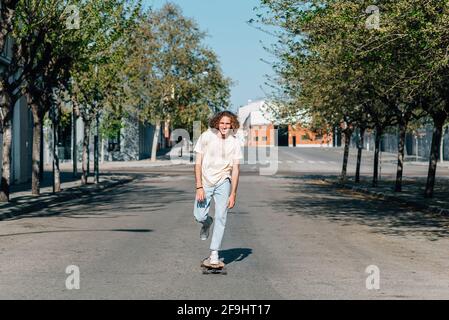 Caucasian red-haired man using a skateboard. He wears casual clothes. He skates down the road of a city surrounded by trees. Sunny summer day with harsh shadows Stock Photo