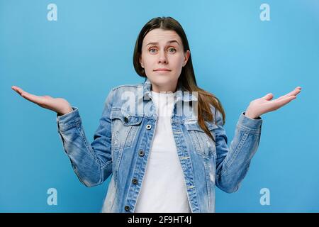 Disgruntled confused annoyed young millennial woman raising hands, indignantly asking reason of failure, wears denim jacket, isolated on blue wall Stock Photo