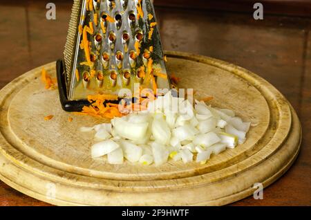 Grated carrots and chopped onions on a cutting board. Raw vegetables and a metal grater on a round wooden cutting board. Cooking homemade food. Health Stock Photo